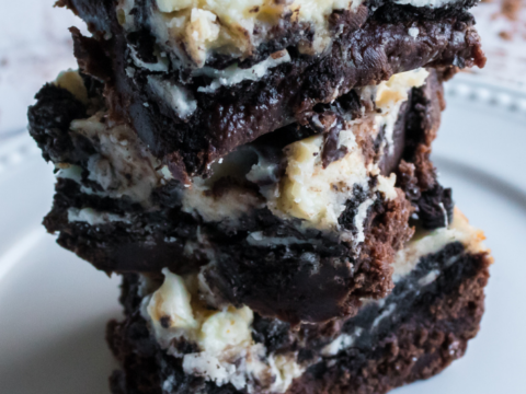 A stack of three OREO Cheesecake Brownies topped with crumbled cookies and creamy frosting sits on a white plate. The brownies have a rich, dark chocolate base with chunky, mixed textures from the toppings.