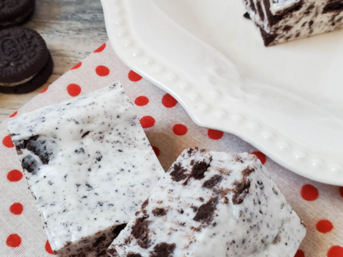 Two squares of OREO White Chocolate Fudge rest on a red polka dot cloth next to a white plate filled with more fudge pieces. Mini chocolate sandwich cookies are scattered across the wooden surface, completing this delectable scene.