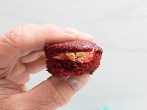 A hand holds a small red velvet cupcake reminiscent of decadent cheesecake brownies, brimming with creamy white and brown filling. The background features a light blue cloth with a checkered pattern, adding a charming touch.