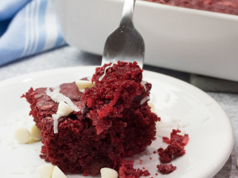 A close-up of a fork holding a bite of Red Velvet Earthquake Cake studded with white chocolate chips on a white plate. In the background, a baking dish overflows with this delightful dessert, while bits of coconut flakes sprinkle the scene.