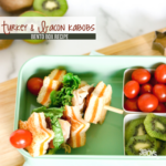 A green bento box features turkey bacon kabobs on skewers, garnished with lettuce and cherry tomatoes. Sliced kiwi and more cherry tomatoes accompany this delightful lunch. The top reads Turkey & Bacon Kabobs Bento Box Recipe.