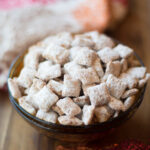 A bowl filled with square-shaped, powdered sugar-coated Thanksgiving puppy chow sits on a wooden surface with colorful fabric in the background.