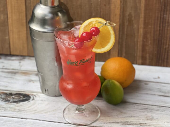 Pink mocktail in a hurricane glass with ice, garnished with an orange slice and cherries, with a cocktail shaker and citrus fruit in the background.
