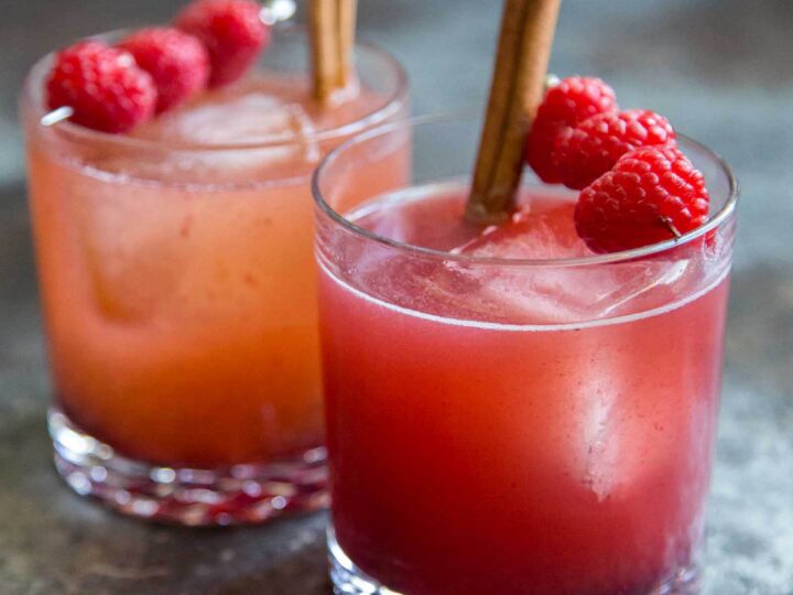 Two berry apple mocktails in short glasses with ice, garnished with fresh raspberries and cinnamon sticks on a dark surface.