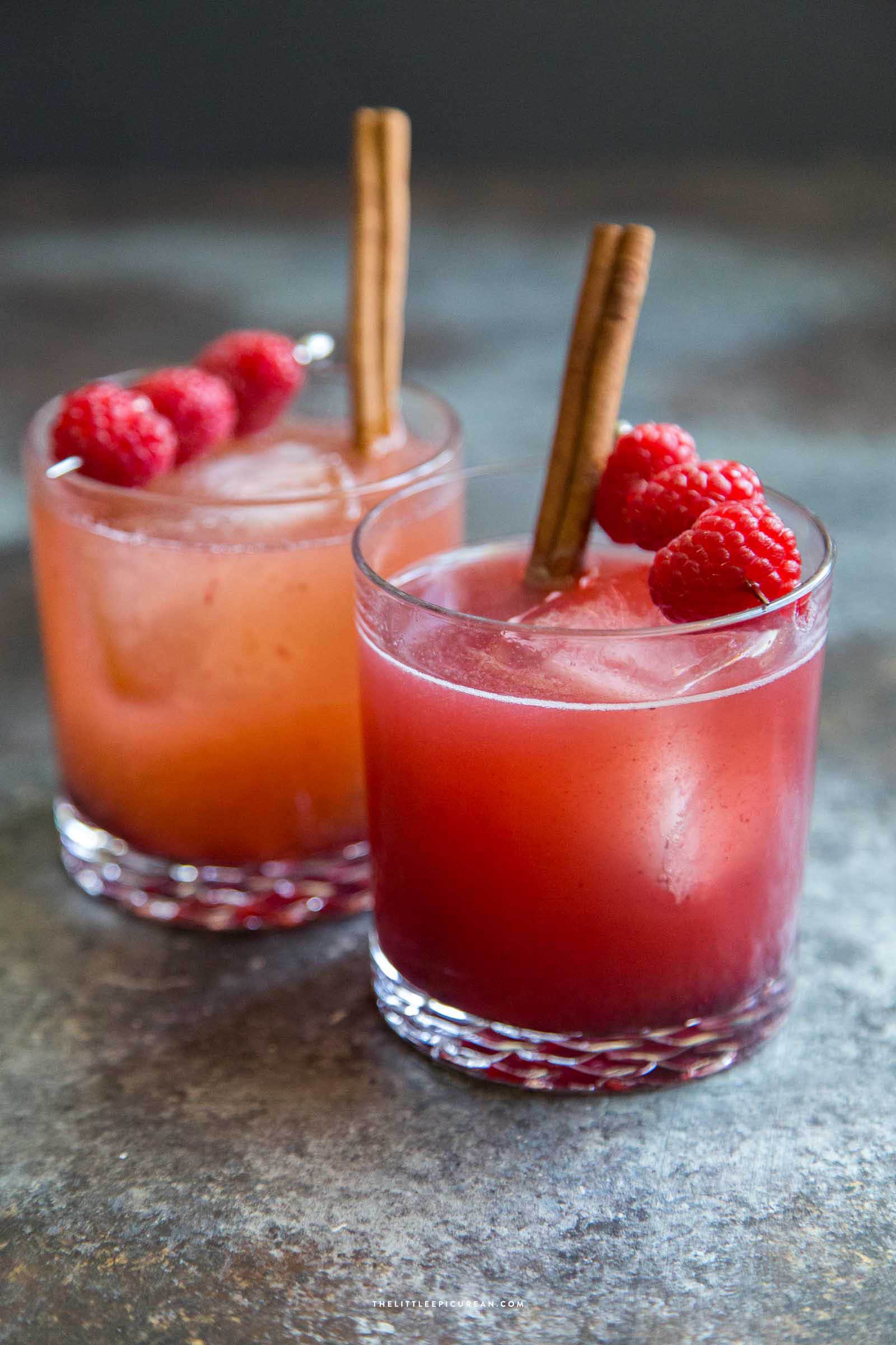 Two berry apple mocktails in short glasses with ice, garnished with fresh raspberries and cinnamon sticks on a dark surface.