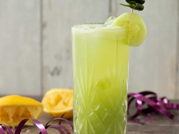 Light green mocktail in a tall glass with ice, garnished with a cucumber slice and curled peel on a skewer, with lemons and ribbon in the background.