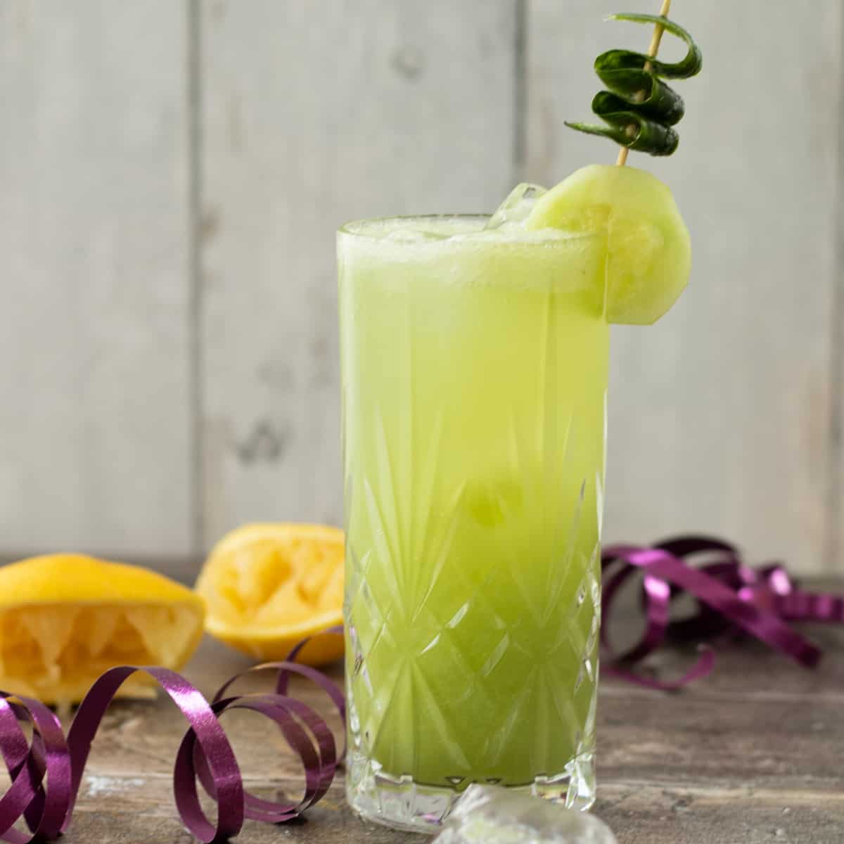 Light green mocktail in a tall glass with ice, garnished with a cucumber slice and curled peel on a skewer, with lemons and ribbon in the background.