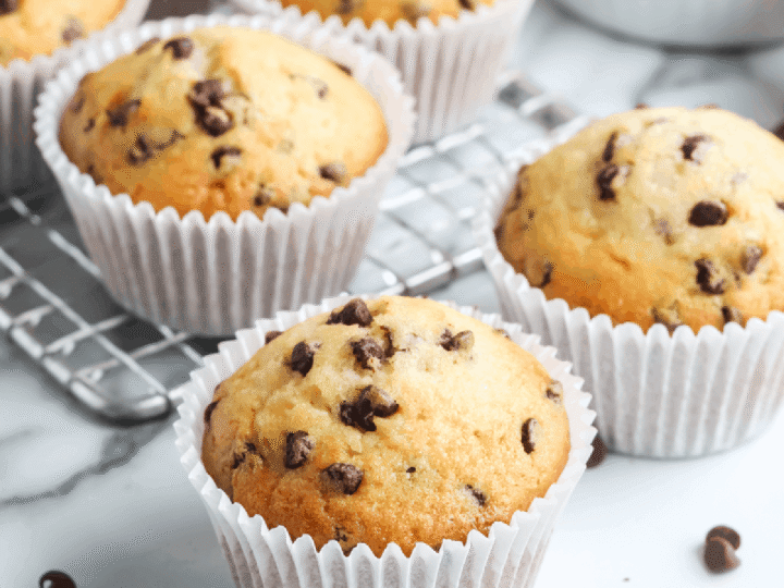 Brunch-perfect chocolate chip mini muffins sit in white paper liners on a cooling rack, with extra chocolate chips scattered across the marble countertop.