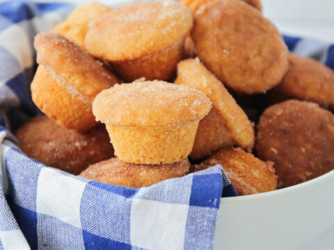 A white bowl filled with sugar-dusted mini muffins is placed on a blue and white checkered cloth, perfect for brunch. The muffins appear golden brown and freshly baked, adding a delicious touch to your morning gathering.