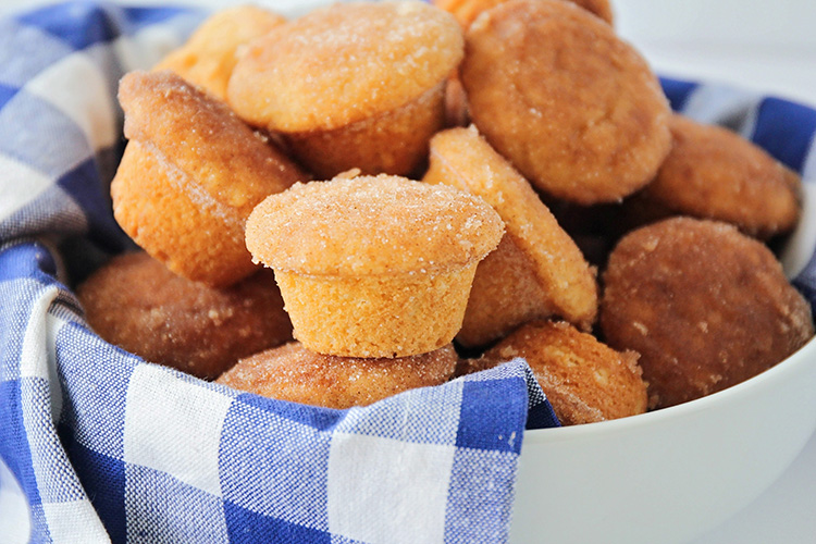 A white bowl filled with sugar-dusted mini muffins is placed on a blue and white checkered cloth, perfect for brunch. The muffins appear golden brown and freshly baked, adding a delicious touch to your morning gathering.