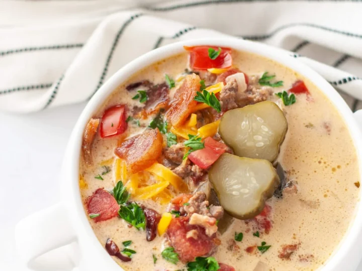 A creamy soup served in a white cup, brimming with flavors of chopped bacon, ground hamburger meat, sliced pickles, red bell peppers, shredded cheese, and parsley. This delightful crockpot meal comes with another cup and a striped cloth in the background.