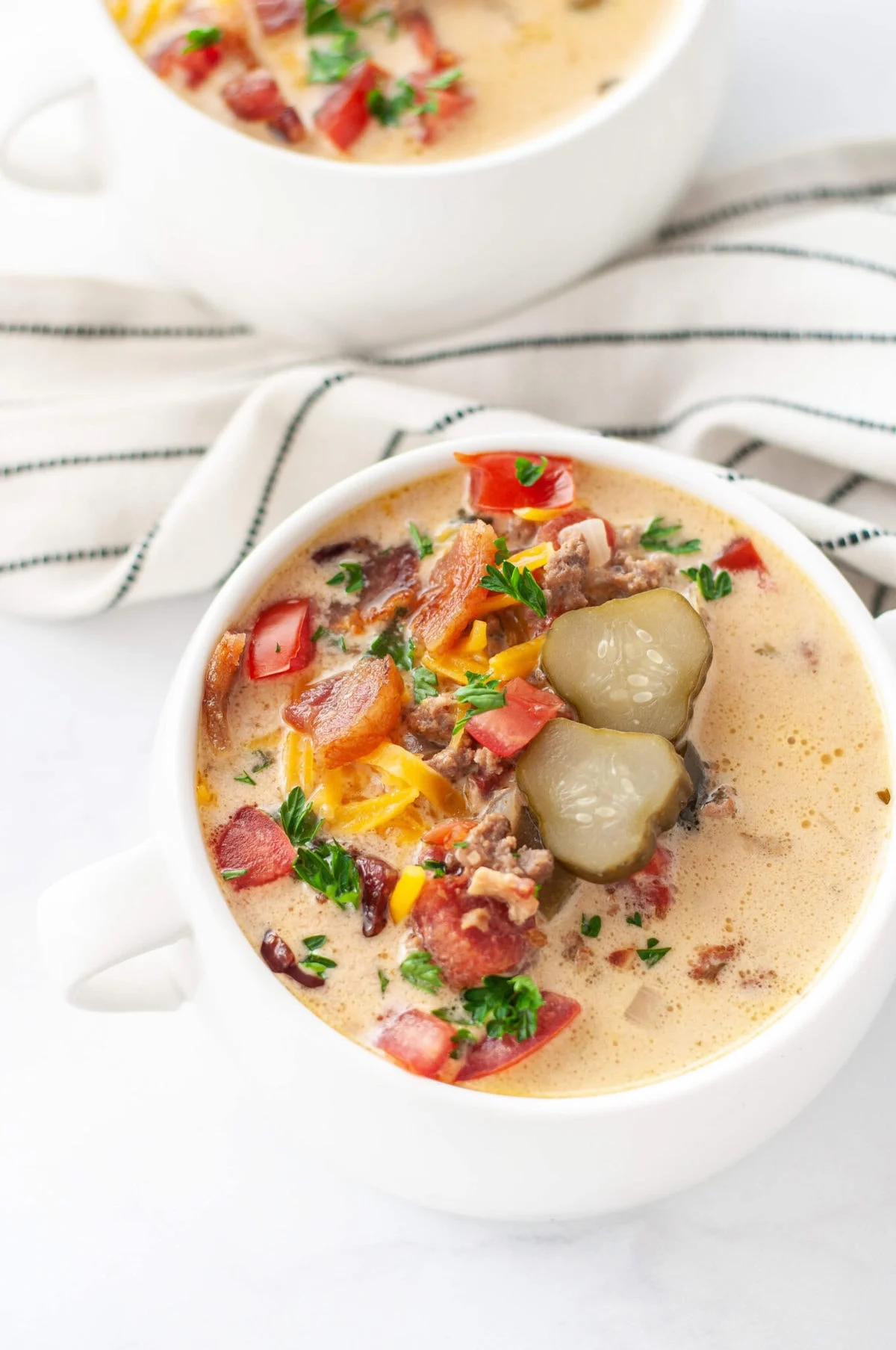 A creamy soup served in a white cup, brimming with flavors of chopped bacon, ground hamburger meat, sliced pickles, red bell peppers, shredded cheese, and parsley. This delightful crockpot meal comes with another cup and a striped cloth in the background.