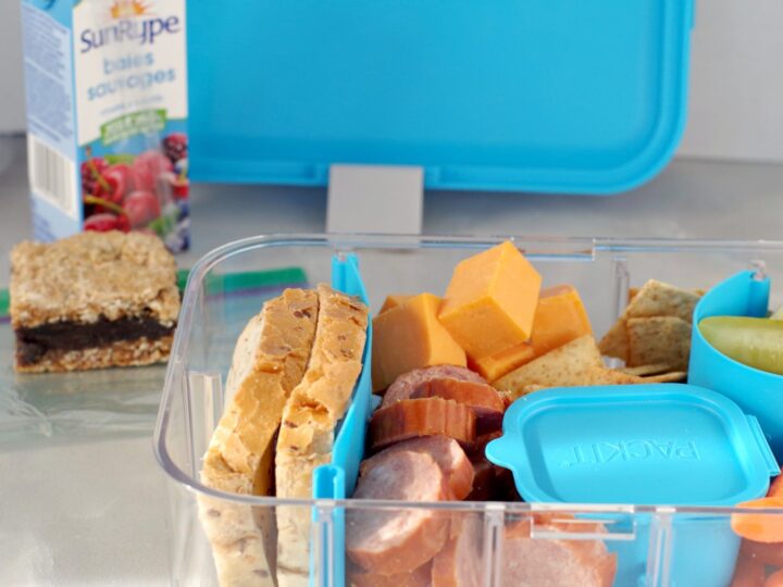 A fun bento box features sliced bread, cheese cubes, sausage, and a small container. Nearby, a juice box and a bar await on the table. The open lunchbox reveals its contents with a blue lid in the background.