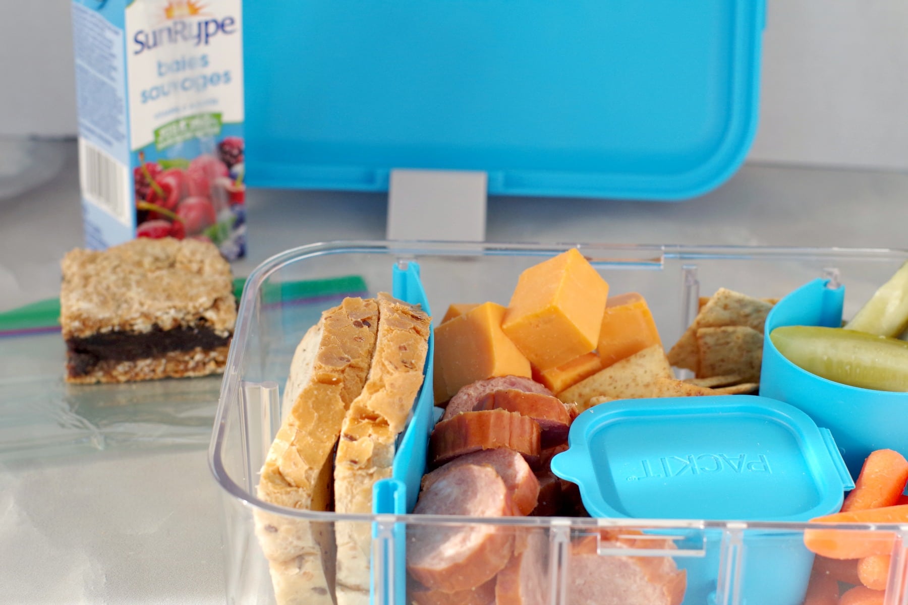 A fun bento box features sliced bread, cheese cubes, sausage, and a small container. Nearby, a juice box and a bar await on the table. The open lunchbox reveals its contents with a blue lid in the background.