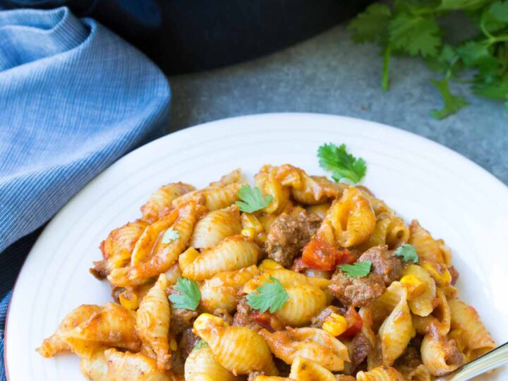 A crockpot meal with pasta shells mixed with ground beef, corn, and diced tomatoes, garnished with cilantro graces the white plate. Nearby, a fork rests alongside a slow cooker and a blue cloth.