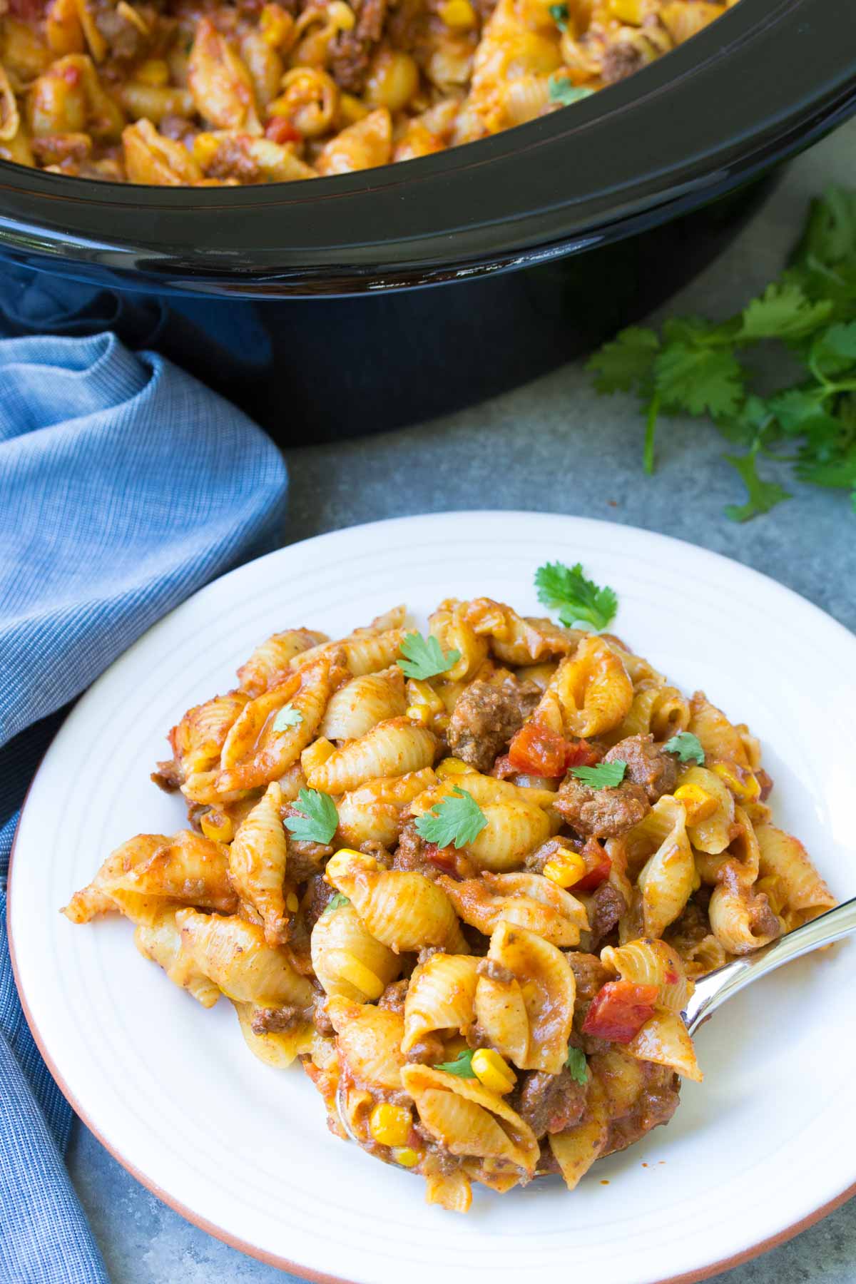 A crockpot meal with pasta shells mixed with ground beef, corn, and diced tomatoes, garnished with cilantro graces the white plate. Nearby, a fork rests alongside a slow cooker and a blue cloth.