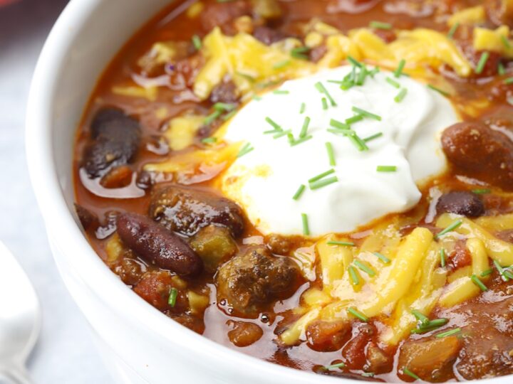 A bowl of savory crockpot chili topped with shredded cheese, a dollop of sour cream, and chopped chives. Brimming with beans and tender chunks of hamburger meat, this hearty meal awaits on a red plate with a spoon at the ready.