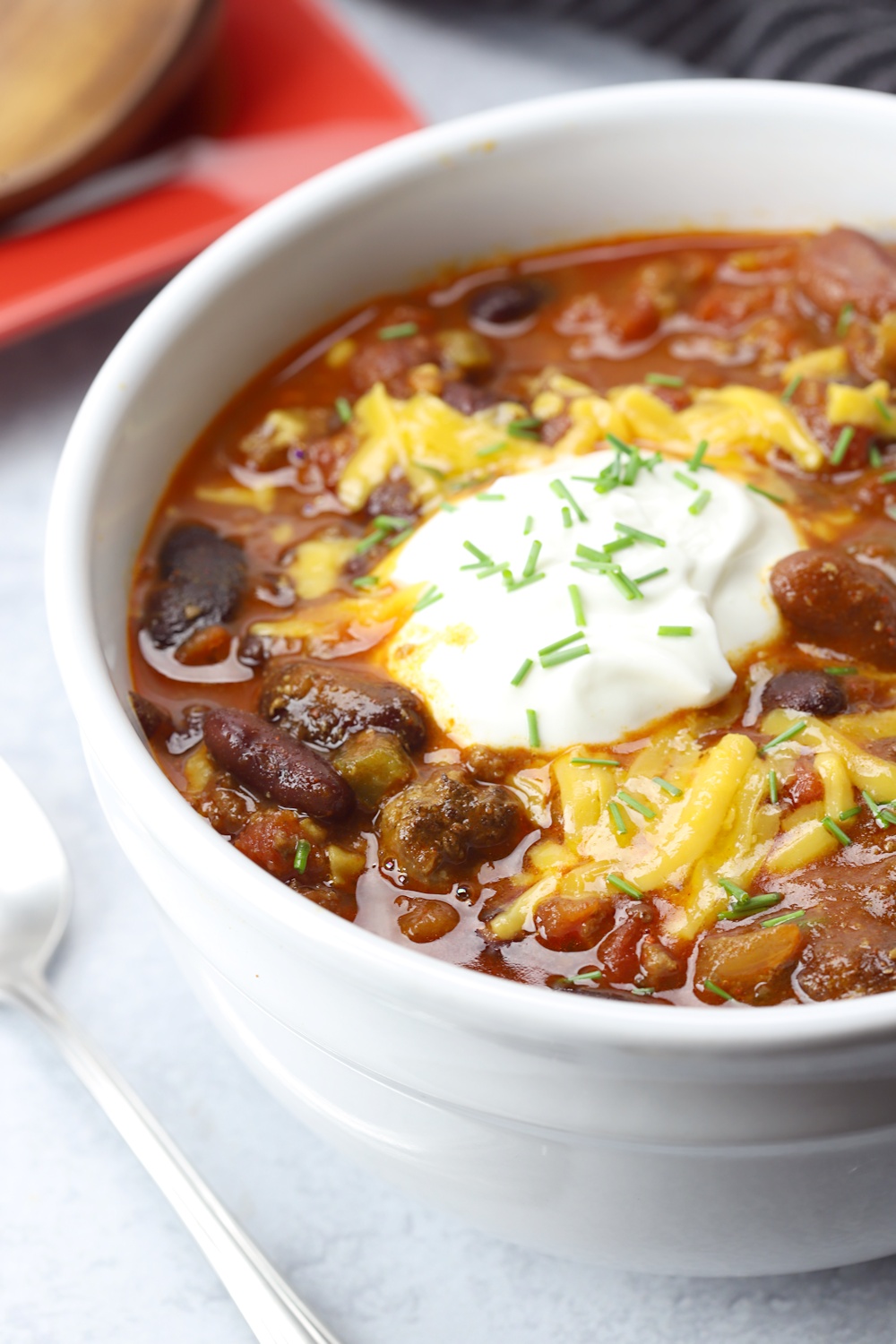 A bowl of savory crockpot chili topped with shredded cheese, a dollop of sour cream, and chopped chives. Brimming with beans and tender chunks of hamburger meat, this hearty meal awaits on a red plate with a spoon at the ready.
