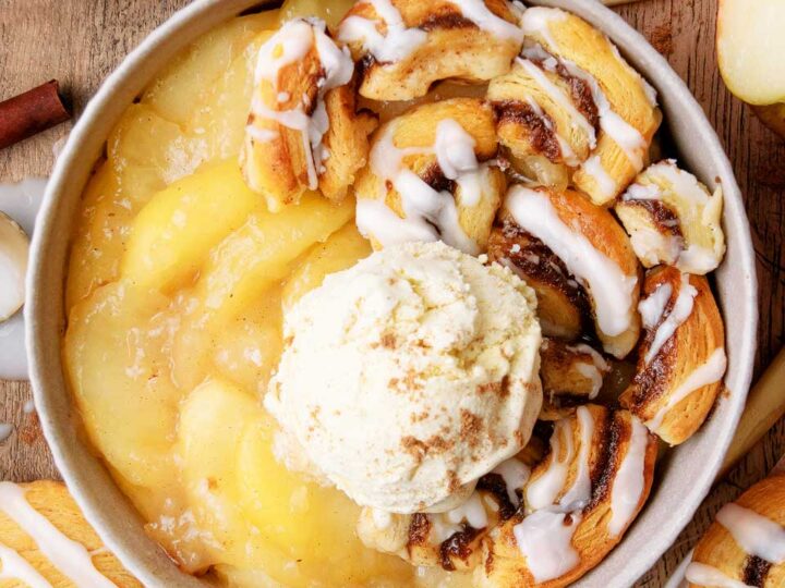 Bowl of warm apple cinnamon dessert with baked apples, cinnamon roll pieces, and vanilla ice cream drizzled with icing on a wooden table.