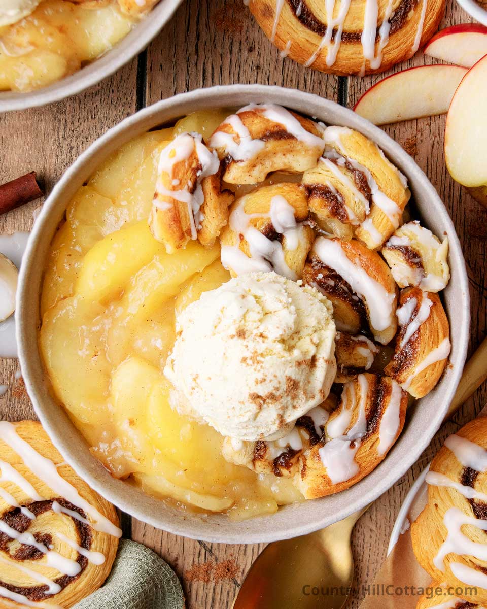 Bowl of warm apple cinnamon dessert with baked apples, cinnamon roll pieces, and vanilla ice cream drizzled with icing on a wooden table.