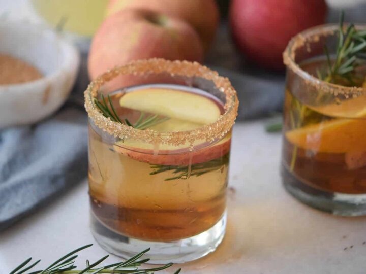 Apple cider mocktail in a short glass with a cinnamon sugar rim, apple slices, and rosemary garnish, with a second drink in the background.