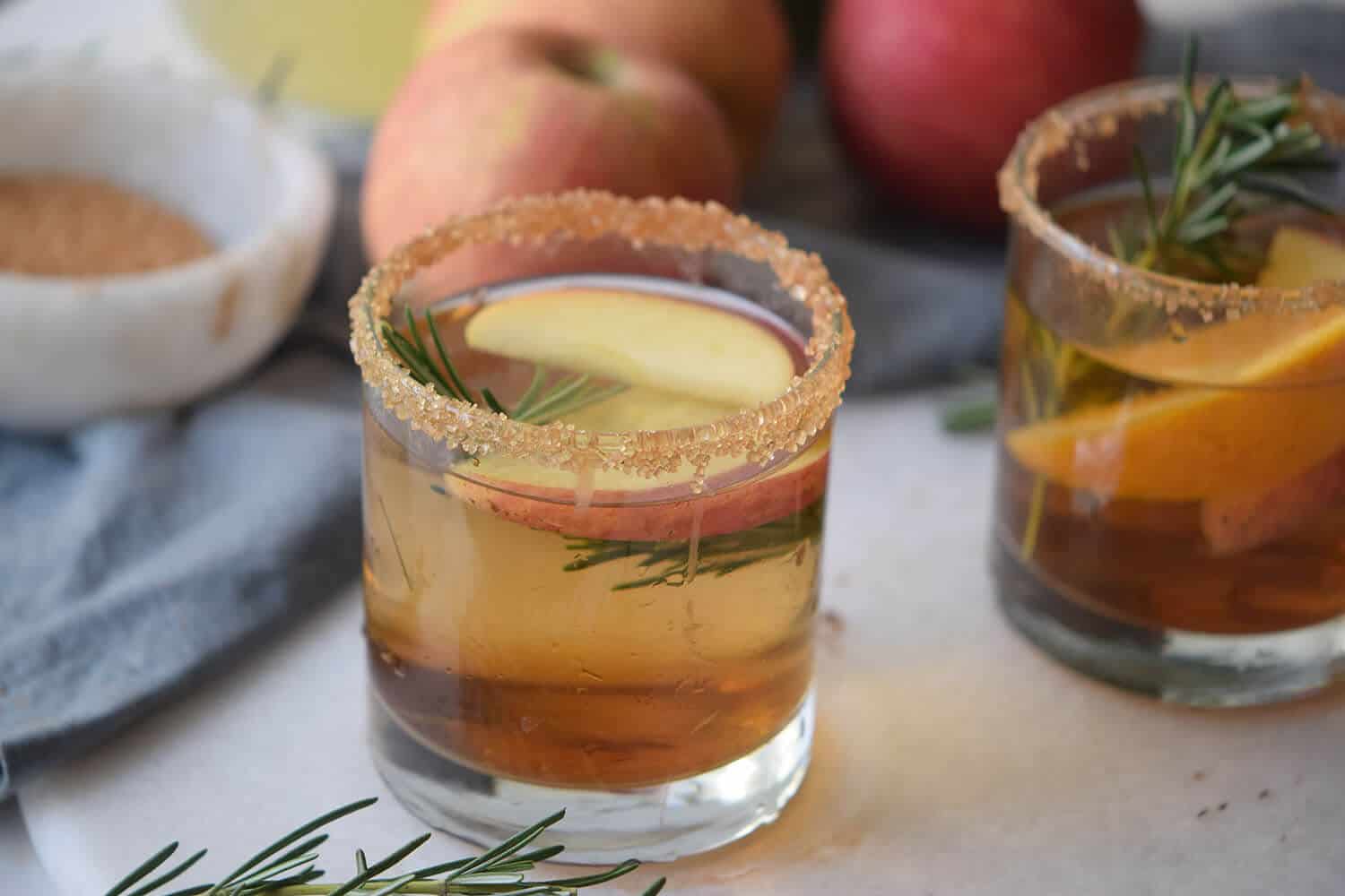 Apple cider mocktail in a short glass with a cinnamon sugar rim, apple slices, and rosemary garnish, with a second drink in the background.