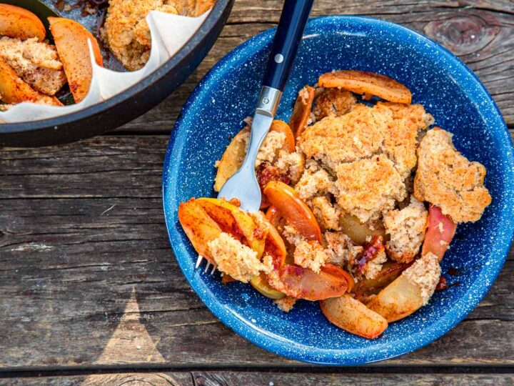 Blue bowl filled with warm apple crisp topped with crumbly streusel and sliced baked apples, served with a fork on a rustic wooden table.