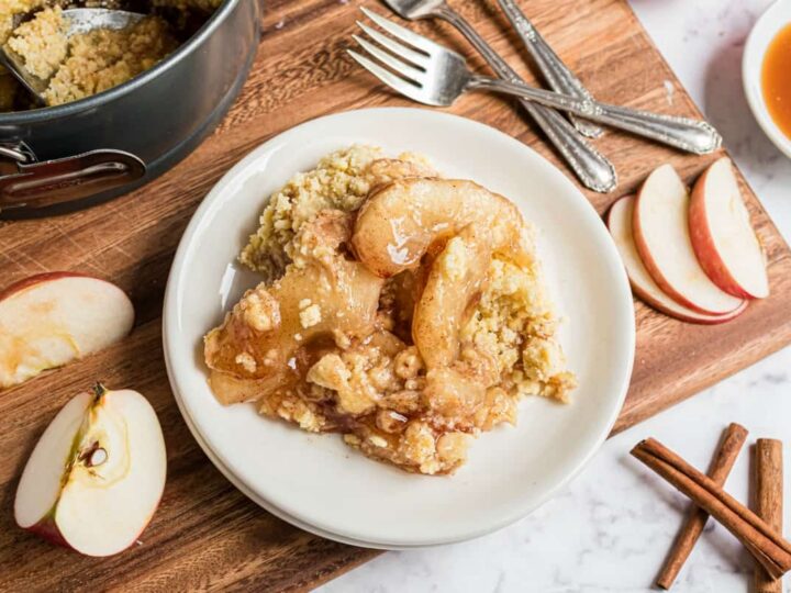 Plate of apple crisp topped with caramel sauce and crumb topping, surrounded by sliced apples, cinnamon sticks, and baking ingredients on a wooden board.