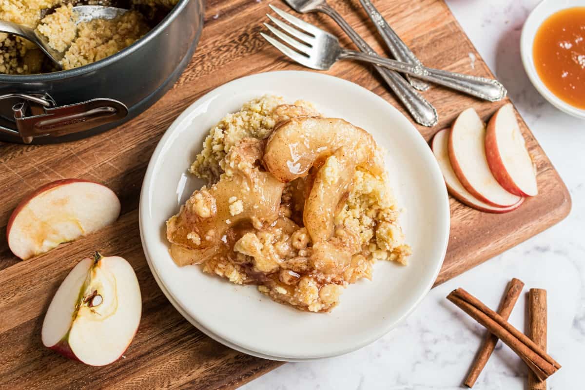 Plate of apple crisp topped with caramel sauce and crumb topping, surrounded by sliced apples, cinnamon sticks, and baking ingredients on a wooden board.