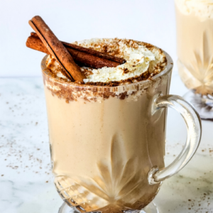 clear glass mug brimming with a creamy pumpkin spice drink recipe, crowned with whipped cream and two cinnamon sticks.