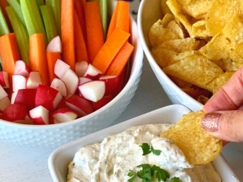 Creamy ranch crack dip in a bowl topped with parsley, served with potato chips, celery, carrots, and radishes for dipping.
