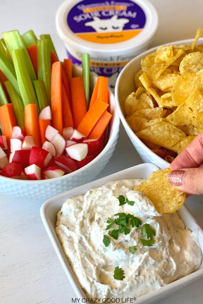 Creamy ranch crack dip in a bowl topped with parsley, served with potato chips, celery, carrots, and radishes for dipping.