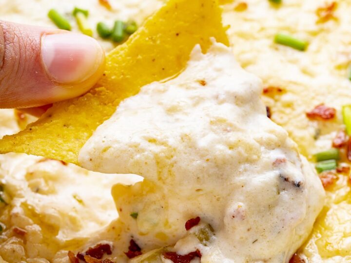 Close-up of a tortilla chip dipped into creamy bacon ranch crack dip with melted cheese and green onions.