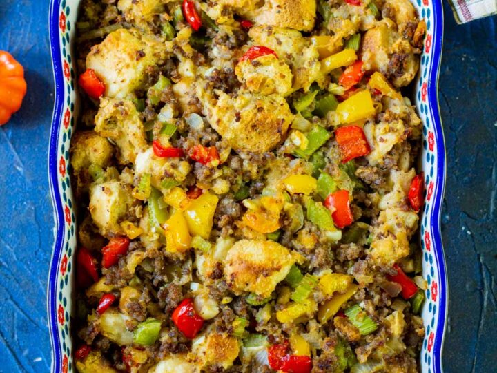 A colorful casserole reminiscent of holiday stuffing recipes sits in a rectangular dish with a floral pattern. It features savory chunks of bread, ground meat, and diced bell peppers and onions. The dish rests on a blue surface, accompanied by a stitched cloth and wooden board.