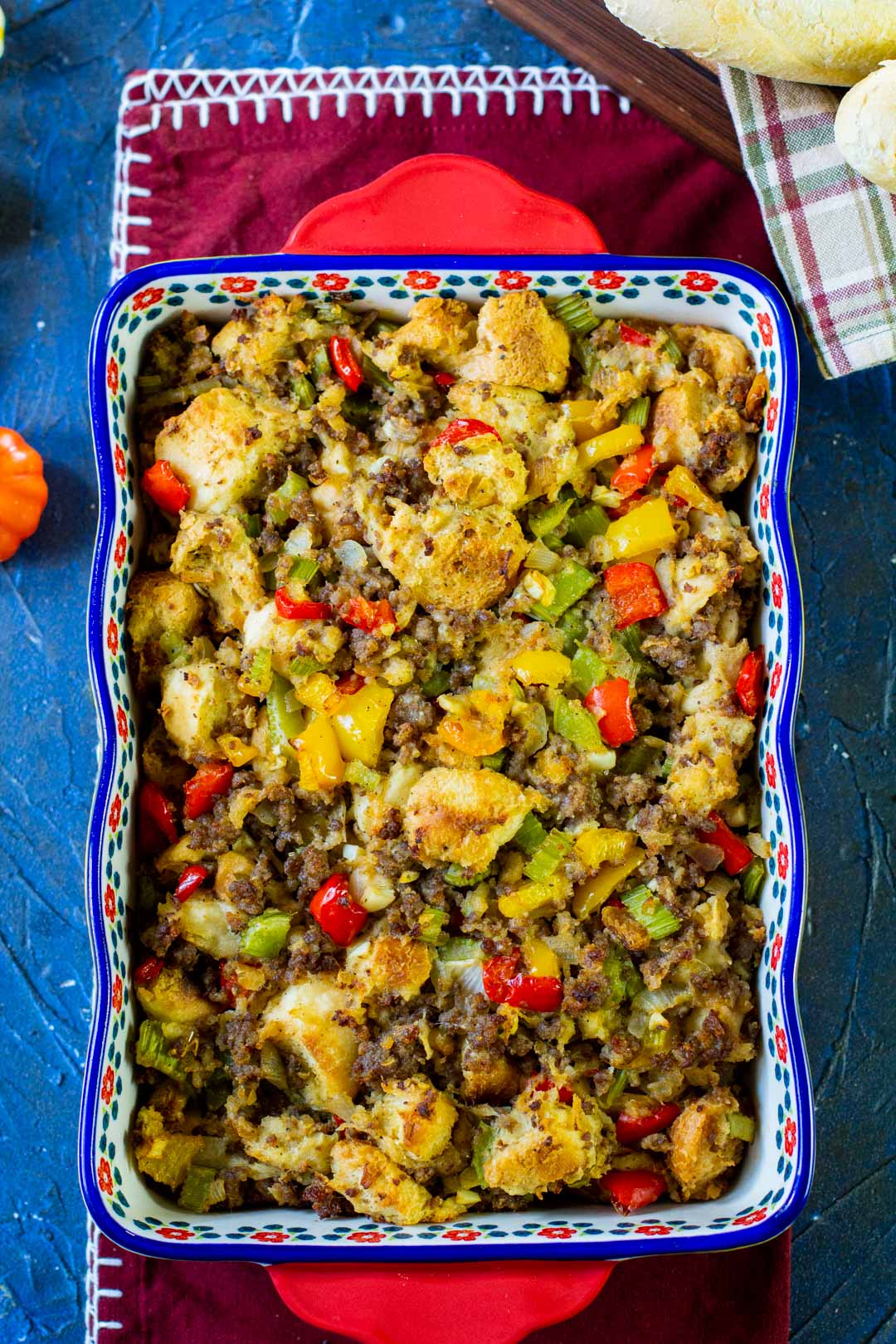 A colorful casserole reminiscent of holiday stuffing recipes sits in a rectangular dish with a floral pattern. It features savory chunks of bread, ground meat, and diced bell peppers and onions. The dish rests on a blue surface, accompanied by a stitched cloth and wooden board.