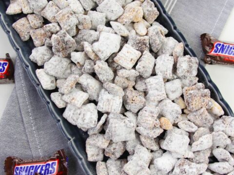 A rectangular black tray brimming with Snickers Puppy Chow is dusted in powdered sugar, perfectly resting on a gray and white striped cloth. Small Snickers bars are artfully scattered around the enticing mix, tempting snack enthusiasts to indulge.