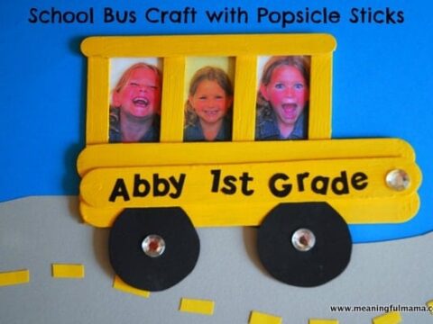 A popsicle stick craft of a yellow school bus with photos of children in the windows. The text says, Abby 1st Grade. The bus has black wheels with shiny centers and is on a blue and gray background.