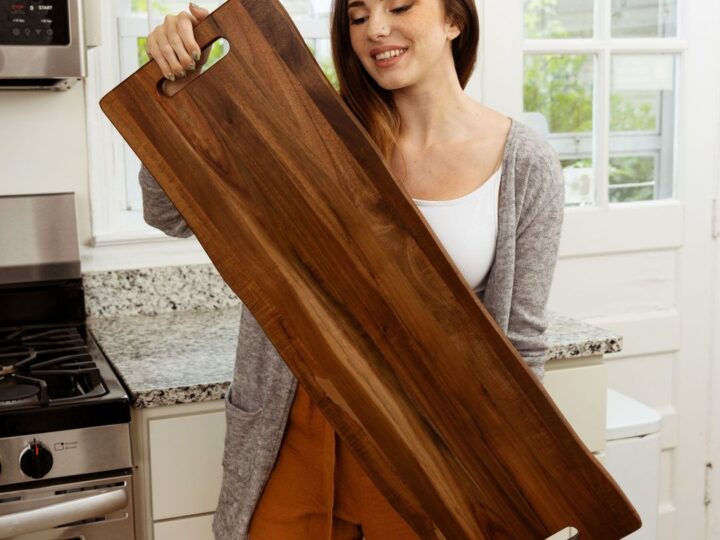 A woman in a kitchen holds a large wooden serving board, perfect for holiday gift ideas. She has long brown hair and is wearing a gray cardigan, white top, and brown shorts. The kitchen features a stove and granite countertops, as sunlight streams in through the window.