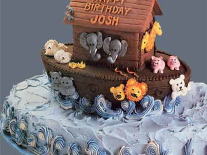 A birthday cake shaped like Noahs Ark, with animal figures like elephants, lions, and pigs on the ark. The cake is decorated with blue icing waves and has the message Happy Birthday Josh on top.