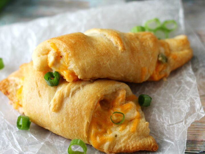 Two golden-brown crescent rolls filled with melted cheese and topped with sliced green onions are placed on crinkled parchment paper.