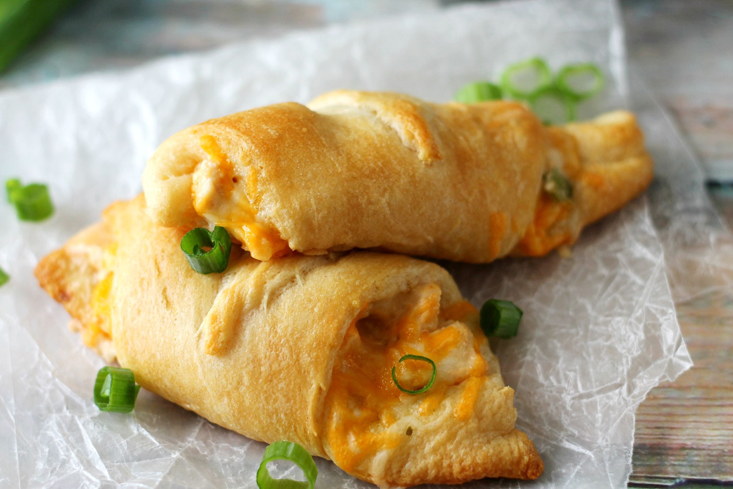 Two golden-brown crescent rolls filled with melted cheese and topped with sliced green onions are placed on crinkled parchment paper.