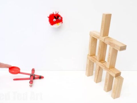 A small red pom-pom with googly eyes is in mid-air, approaching a structure made of wooden blocks. Below, a person flicks a red catapult, seemingly launching the pom-pom. The setup resembles a playful game.