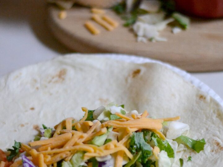 A soft tortilla topped with shredded cheese, lettuce, cabbage, and cilantro. In the background, bowls of additional cheese and salad ingredients sit on a wooden board.