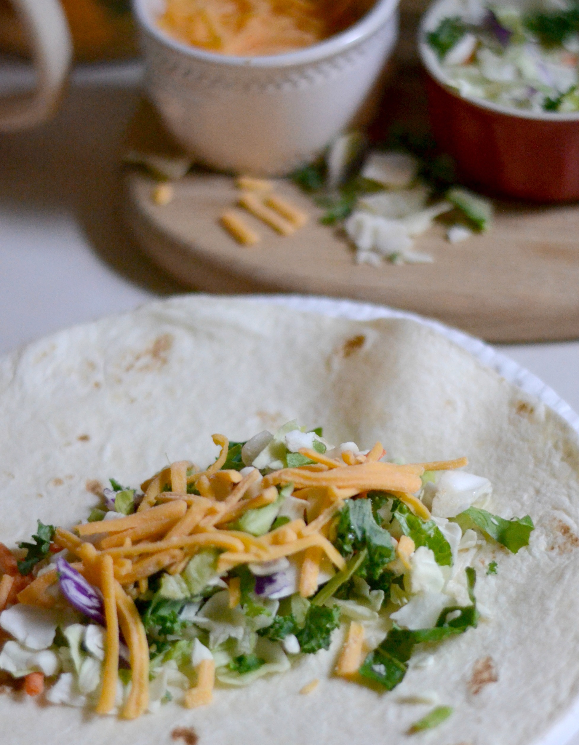 A soft tortilla topped with shredded cheese, lettuce, cabbage, and cilantro. In the background, bowls of additional cheese and salad ingredients sit on a wooden board.