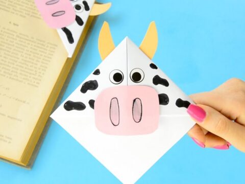 A hand holding a cow-themed origami bookmark, featuring a pink snout, googly eyes, and yellow horns. Its placed on a blue background next to an open book. The design is playful and creative.