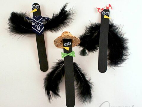Three craft stick crows with black feather wings. Each has googly eyes and a yellow beak. One wears a red bowtie and kerchief, another a green bowtie and straw hat, the third a blue kerchief. They are on a white background.