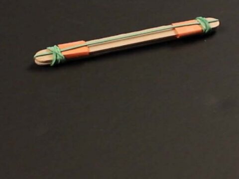 A homemade slingshot made from two popsicle sticks, two rubber bands, and a small piece of paper is placed on a black surface.