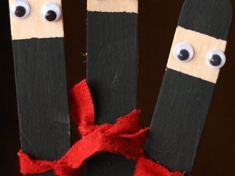 Craft stick ninja ornaments with black painted faces, white googly eyes, and red fabric bands. The image features three sticks arranged closely together. Text reads Craft Stick Ninja Ornaments.