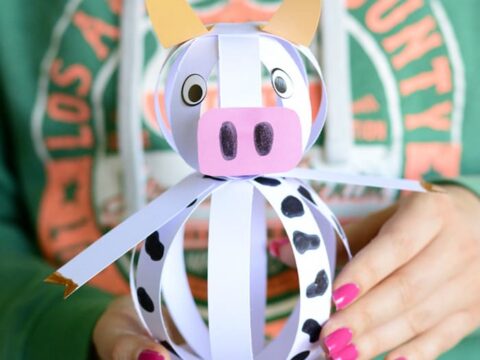 A person holds a paper craft shaped like a cow made from strips of white paper with black spots, featuring exaggerated eyes and ears. The person is wearing a green shirt with an orange and white circular design.