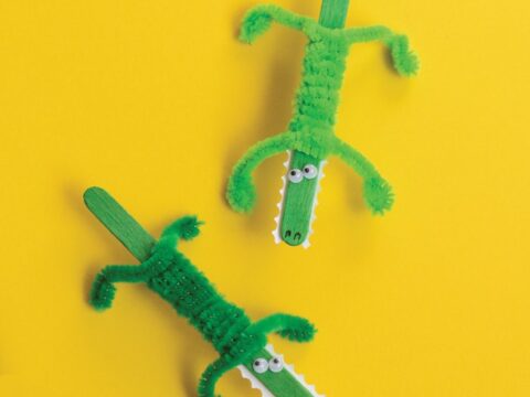 Two green alligator crafts made from craft sticks, pipe cleaners, and googly eyes on a yellow background.